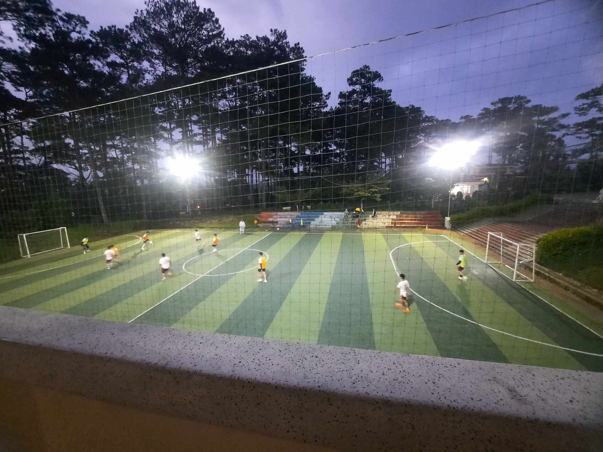 Campo de fútbol reducido iluminado por focos al anochecer, con jugadores sobre el césped